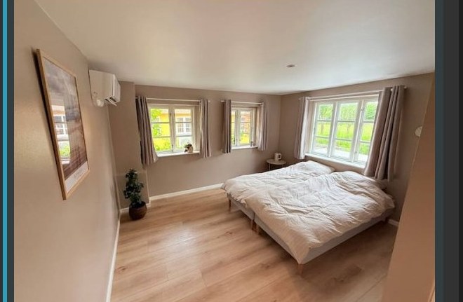 The Dueodde Room — a bright bedroom with a double bed in white linen, light hardwood floors, and three windows opening onto the garden.
