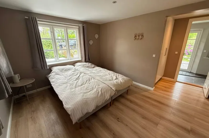 The Dueodde room from inside — oak floor, double bed, and three windows letting in the morning light, with a doorway out to the entrance hall.