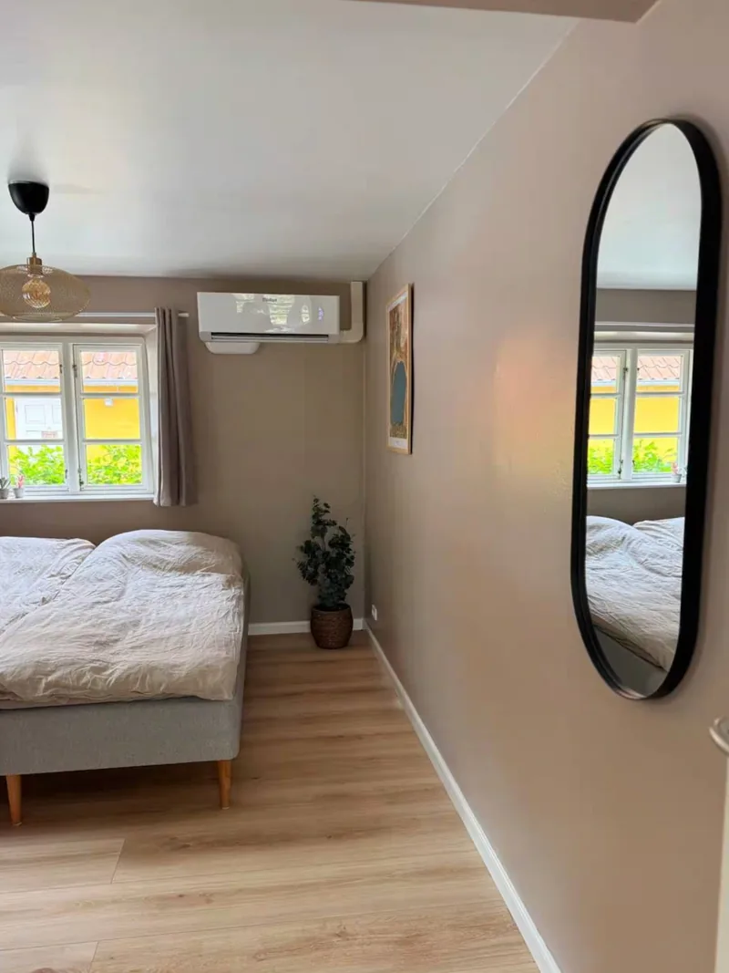 The Opalsøen room from inside — a bedroom with a double bed, an oval black-framed mirror, a pendant lamp, and a window onto Svaneke's yellow façades.