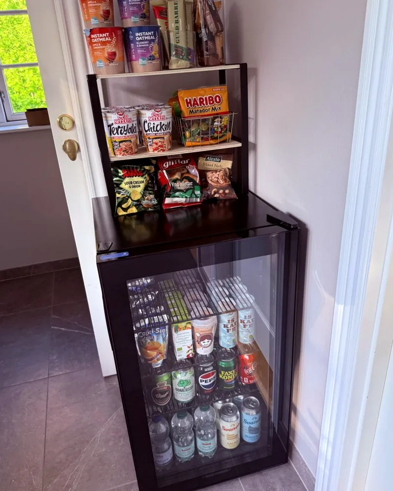 The Trust Bar — a self-serve pantry with a small fridge and a shelf of snacks, soft drinks, instant noodles, and Danish sweets, free for all guests.