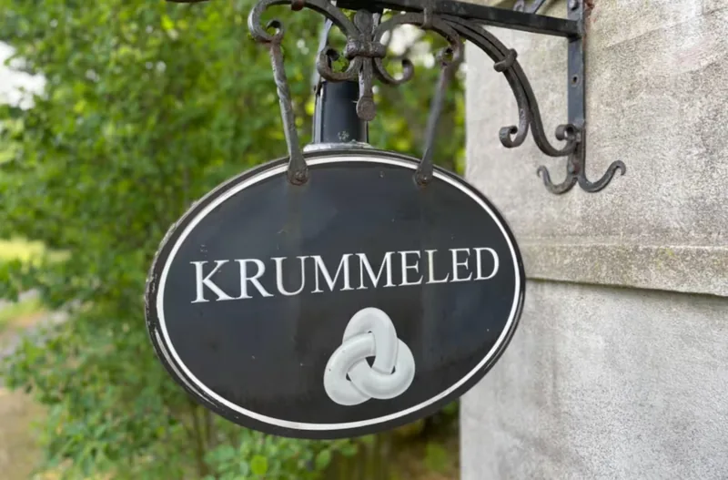 Krummeled's house sign — a black enamel oval with white lettering and a knot symbol, hung in a wrought-iron bracket.