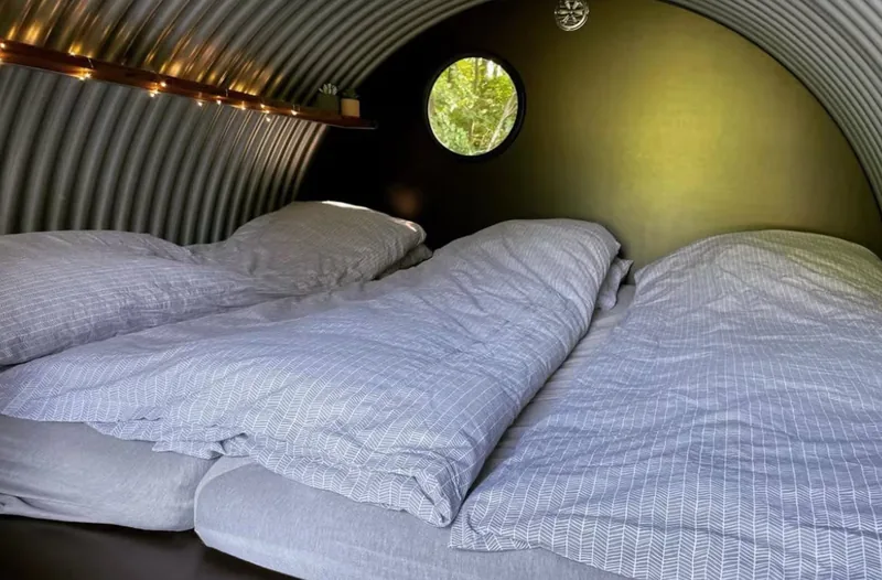 Inside the shelter — striped grey bed linen on the double bed, a small porthole onto green leaves.