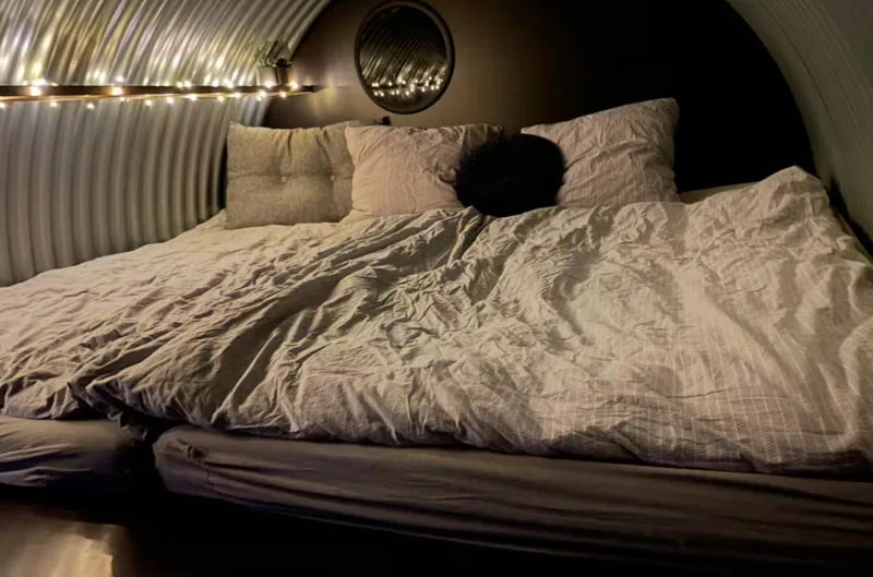 The shelter from inside, the bed made up in grey linen, string lights along the corrugated wall, and a round mirror above.