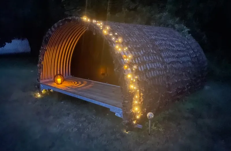 The Luxury Shelter at night — the corrugated arch draped in string lights under dark trees, a paper lantern glowing on the deck.