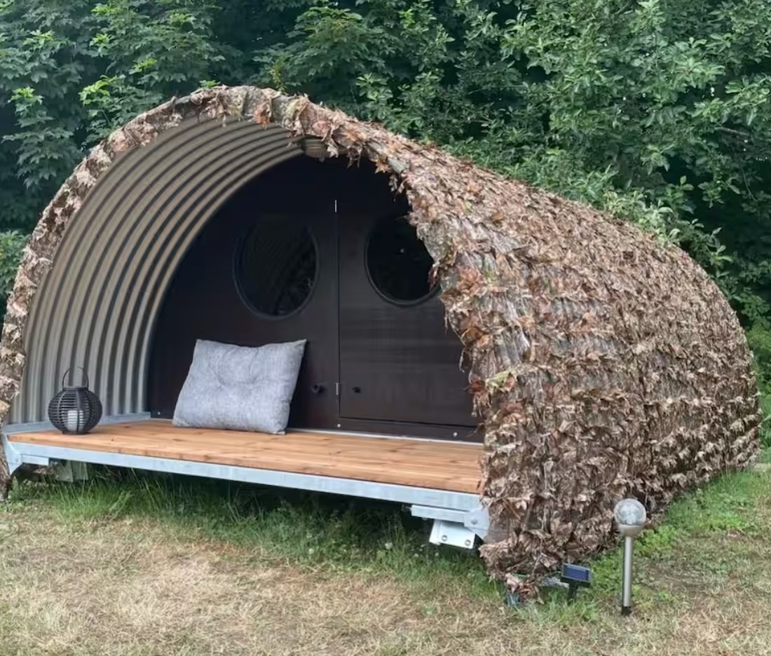The Luxury Shelter — a curved metal cabin with a leaf-thatched cover, porthole windows, and a wooden deck under the trees.
